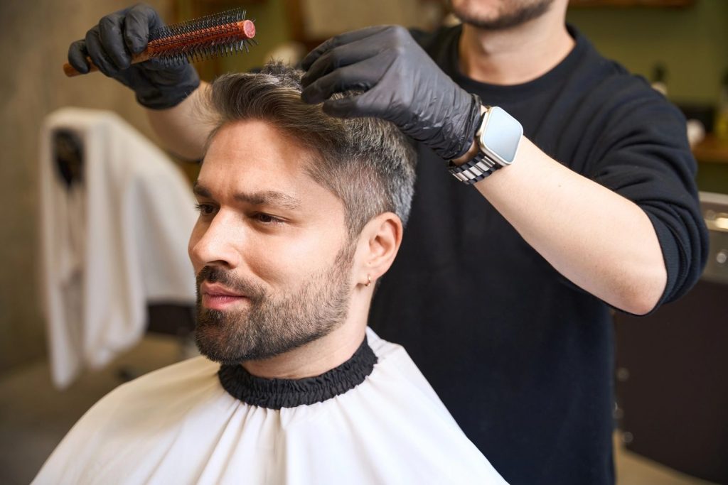 Stylist expertly grooming a man's salt-and-pepper hair using a brush in a Stafford salon specializing in men's hair replacement systems.