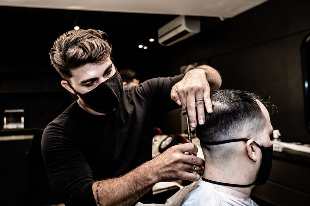 Modern barber shop scene: a masked barber using a straight razor to detail a client's stylish faded haircut. 